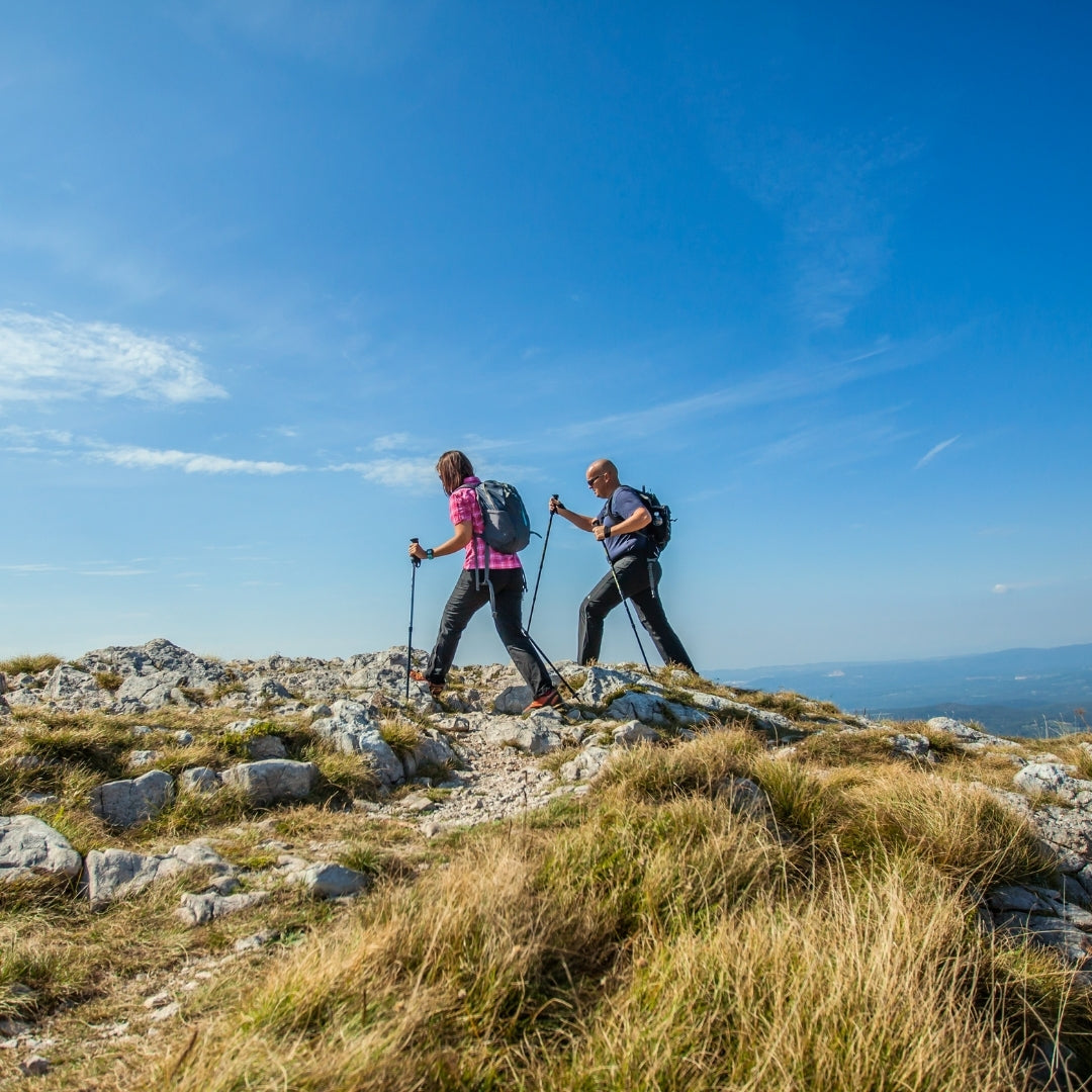 Two hikers with backpacks and trekking poles walk along a rocky grassy mountain ridge under a bright blue sky.