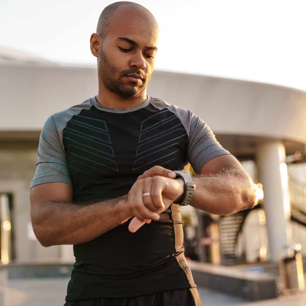 A man in a black and gray athletic shirt checks the time on a smartwatch outdoors in a sunny, modern setting.