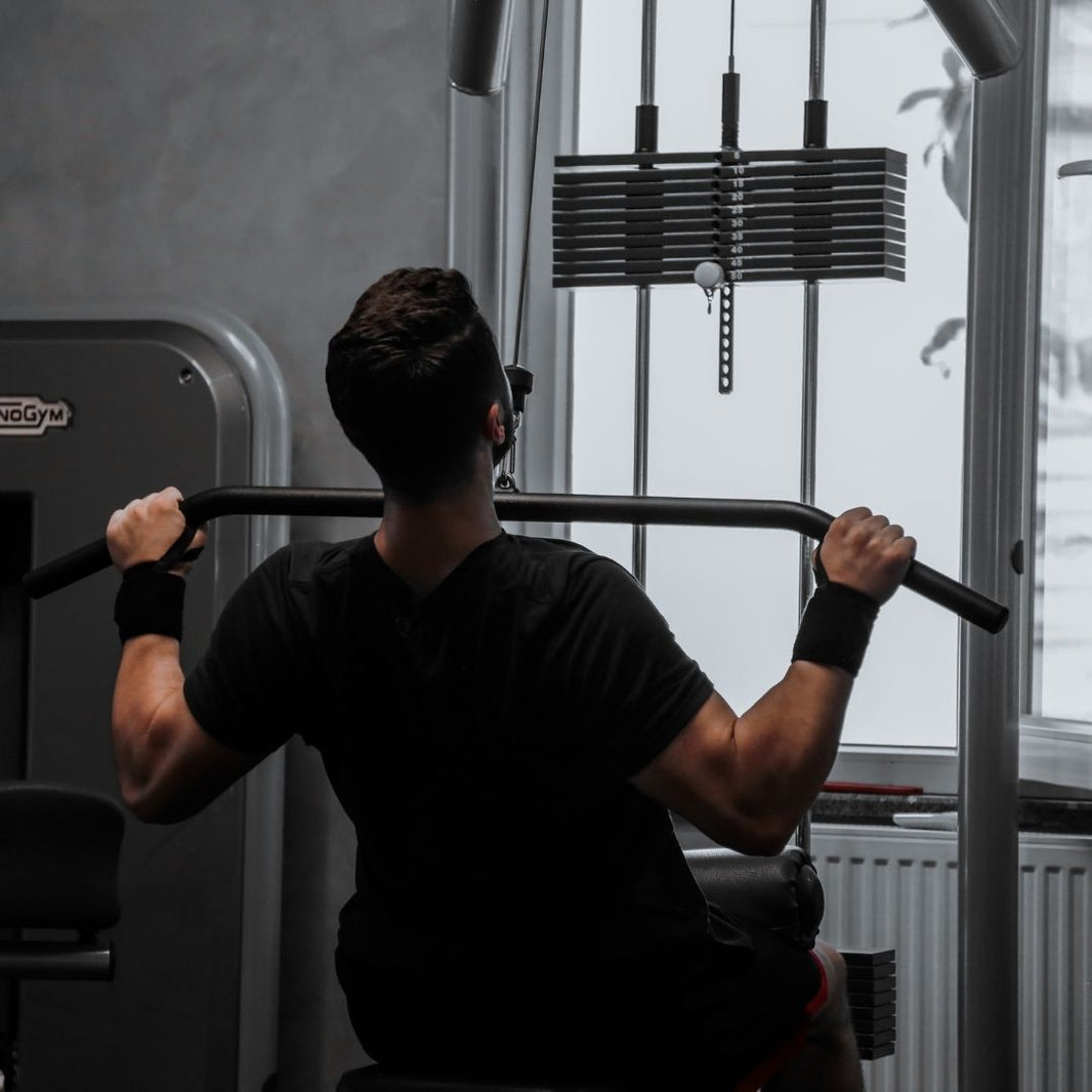A man viewed from behind using a cable pulley exercise machine in a gym, wearing a black t-shirt and wrist wraps.