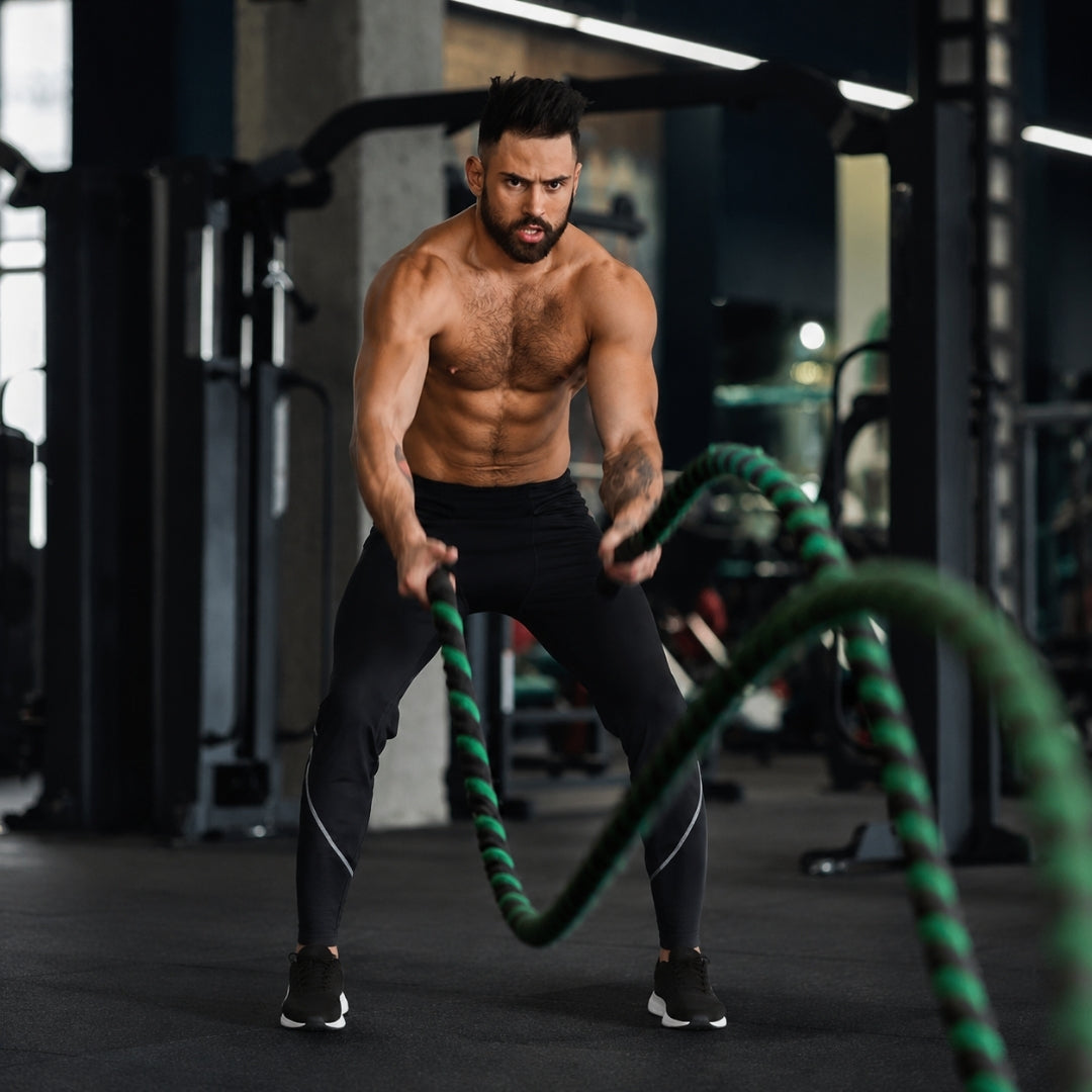 A shirtless, muscular man in a gym wearing black athletic pants and black sneakers, performing battle rope exercises with a green and black battle rope.