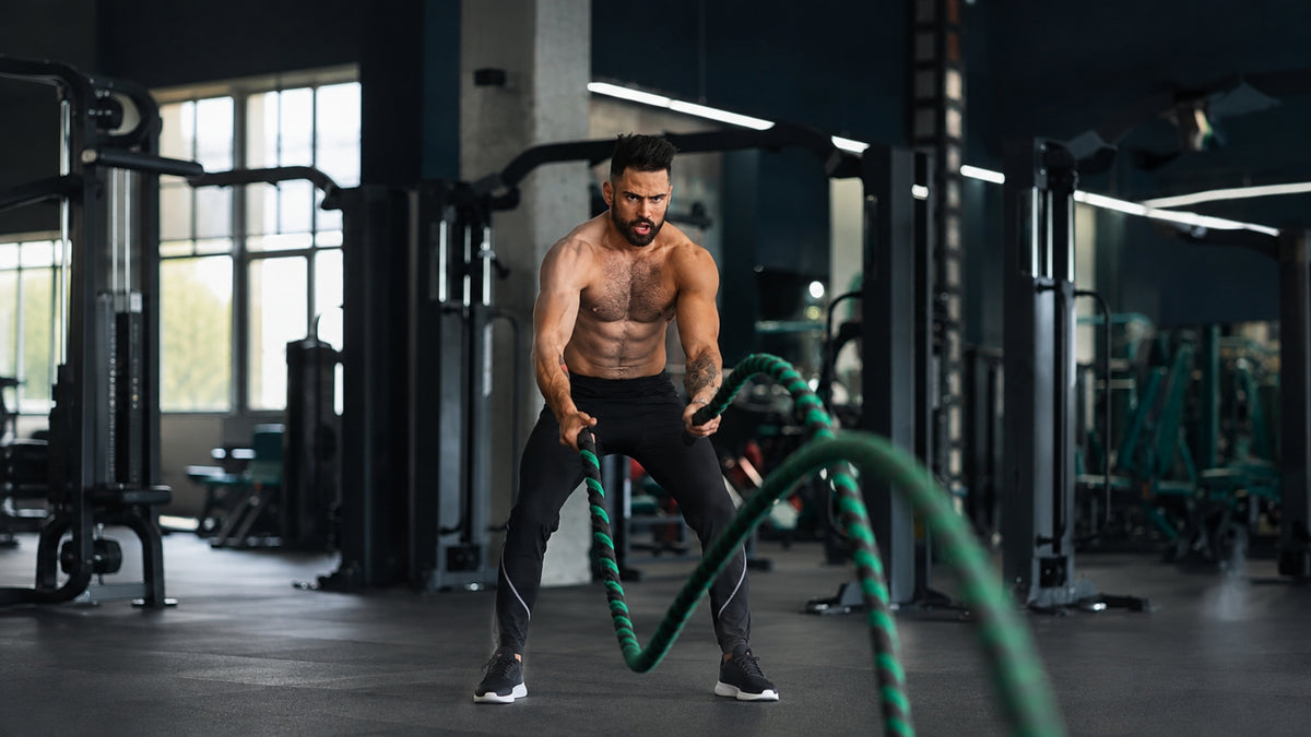 A shirtless, muscular man in a gym wearing black athletic pants and sneakers, holding and battle-roping in a dark gym with exercise equipment in the background.